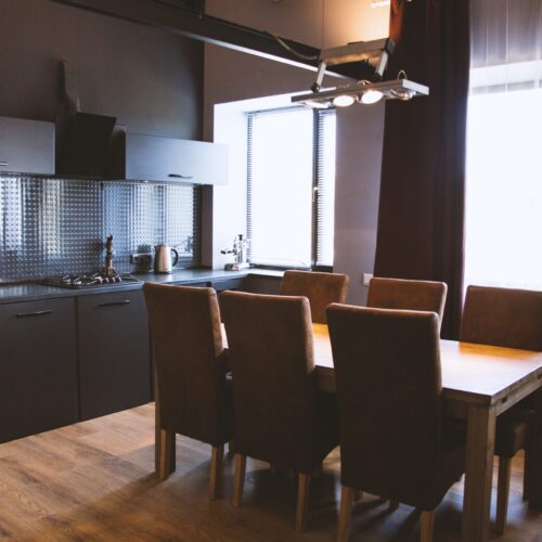 A wide shot of a wooden table with wooden chairs near window curtains  in a kitchen with black interior